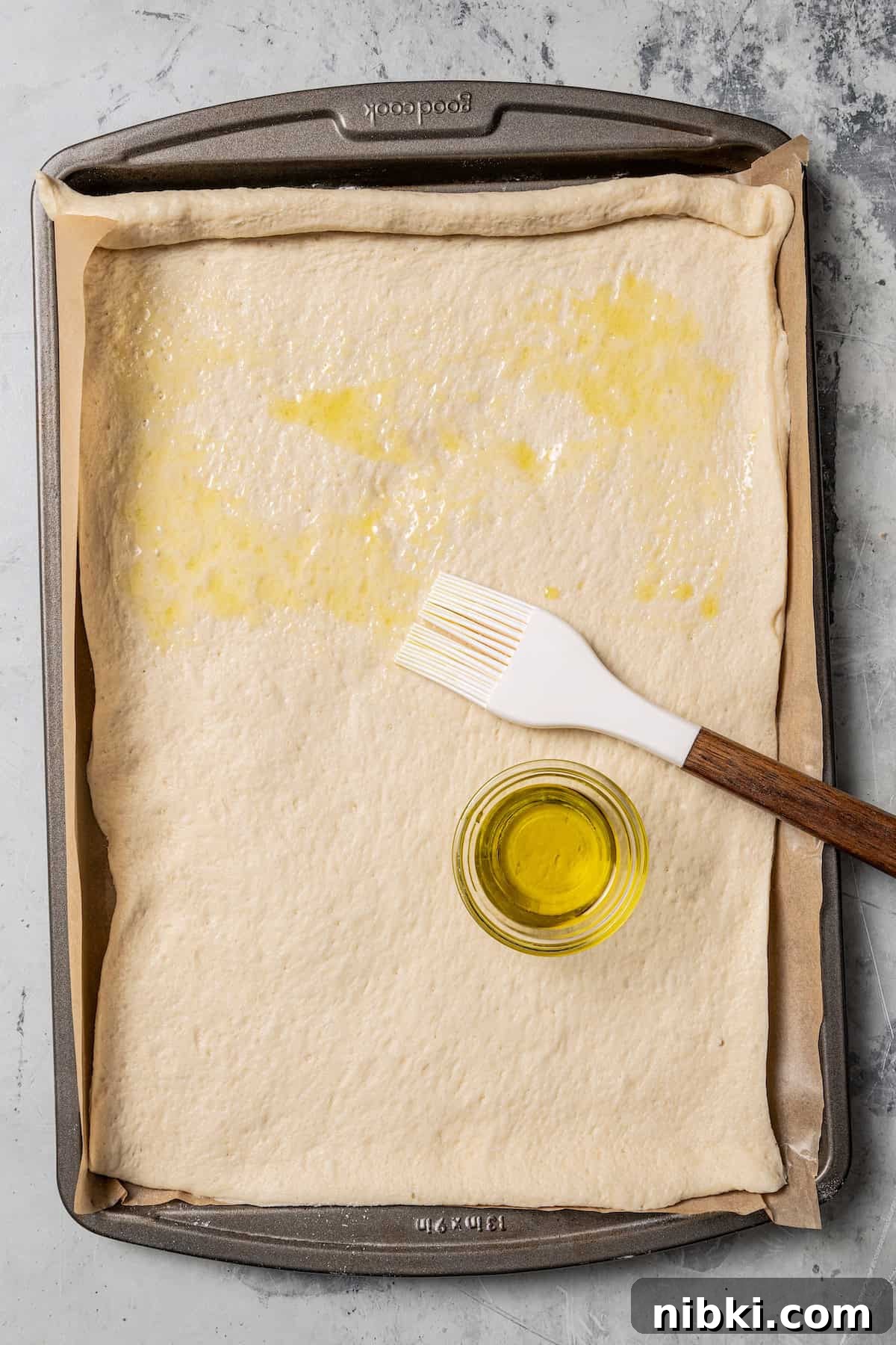 Cheesy pizza rolls are the best appetizer! Flaky dough gets rolled up with tangy tomato sauce, melty mozzarella, and pepperoni. Overhead view of rolled out pizza dough being brushed with olive oil on a parchment-lined baking sheet.