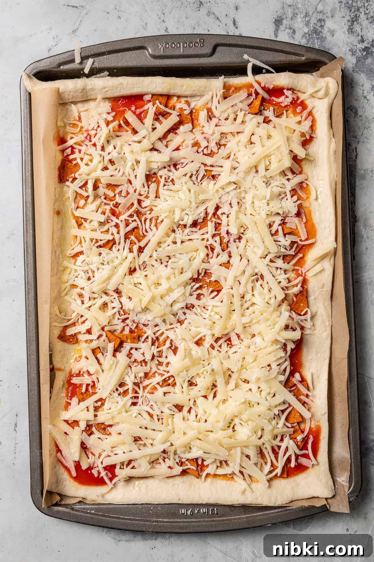 Cheesy pizza rolls are the best appetizer! Flaky dough gets rolled up with tangy tomato sauce, melty mozzarella, and pepperoni. Overhead view of rolled out pizza dough topped with sauce and shredded cheese on a parchment-lined baking sheet.