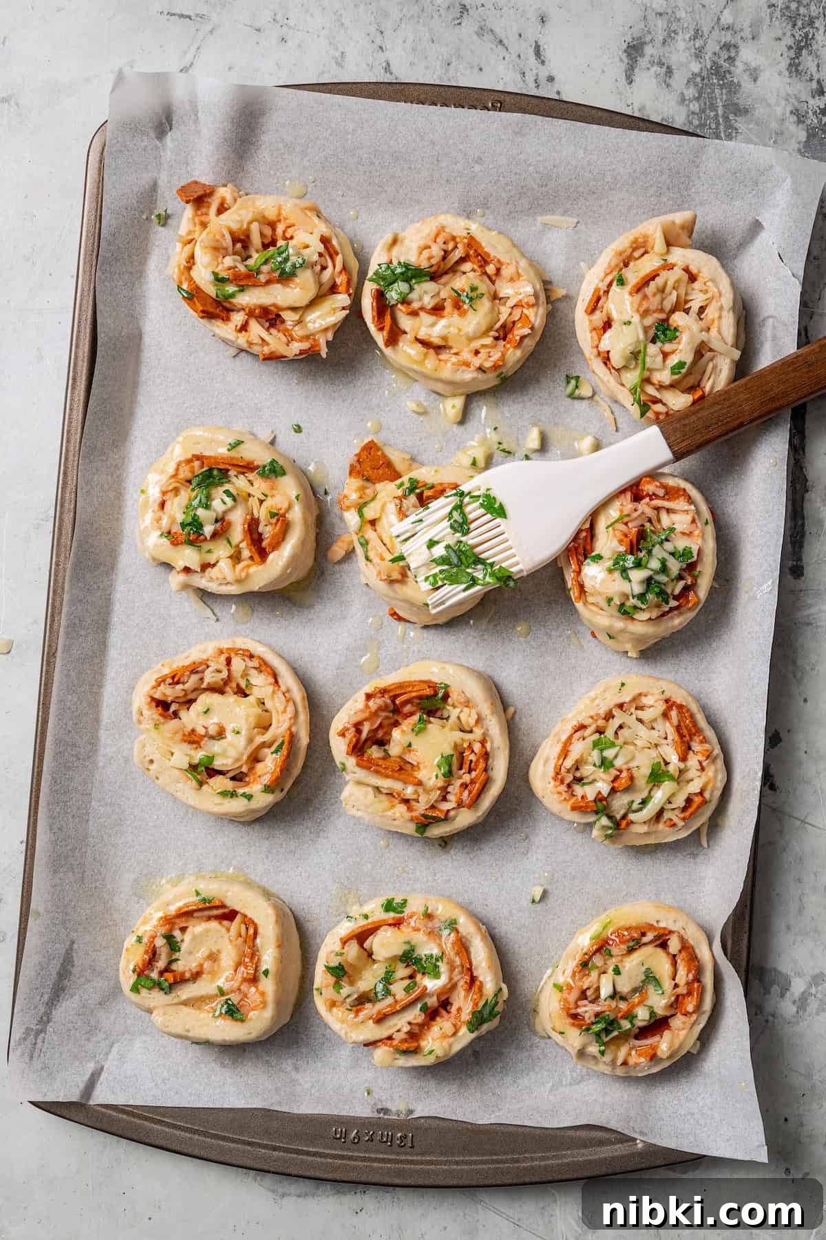 Cheesy pizza rolls are the easiest and best appetizer, made with flaky dough, tangy tomato sauce, melty mozzarella, and pepperoni. Overhead view of pepperoni pizza rolls being brushed with garlic butter on parchment-lined baking sheet.