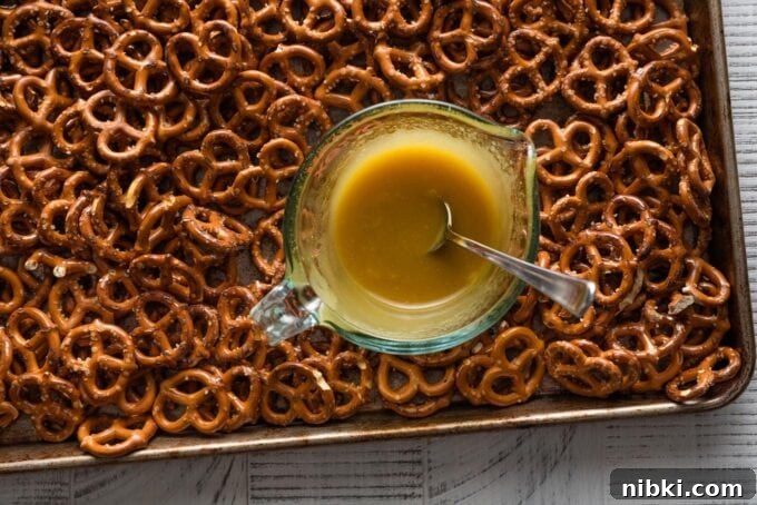 Landscape image of a large baking sheet spread with miniature pretzels and a small glass liquid measuring cup with a honey mustard seasoning blend mixed together in the middle, ready to pour over.