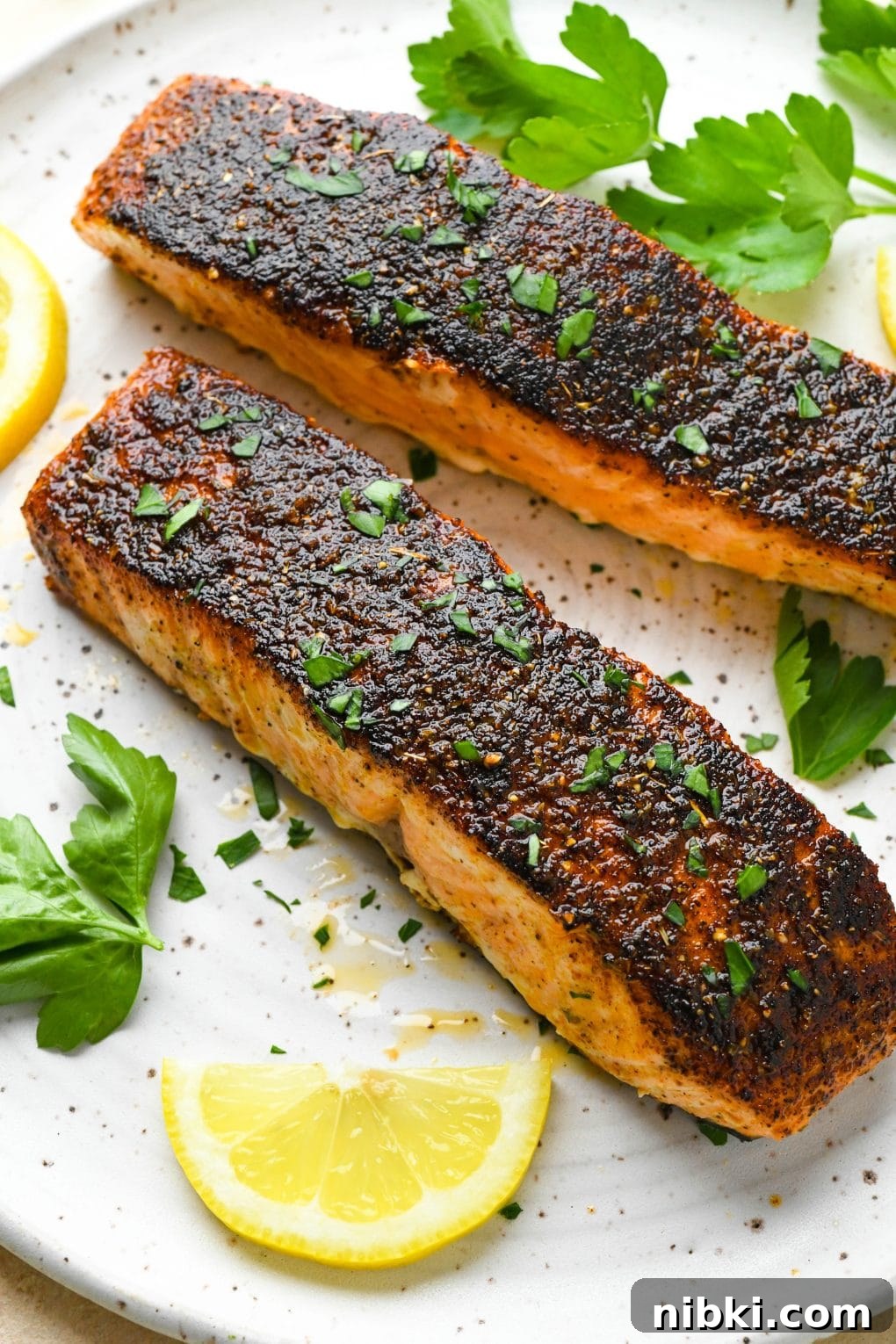 Two blackened salmon filets on a white speckled plate, surrounded by fresh herbs and lemon wedges.
