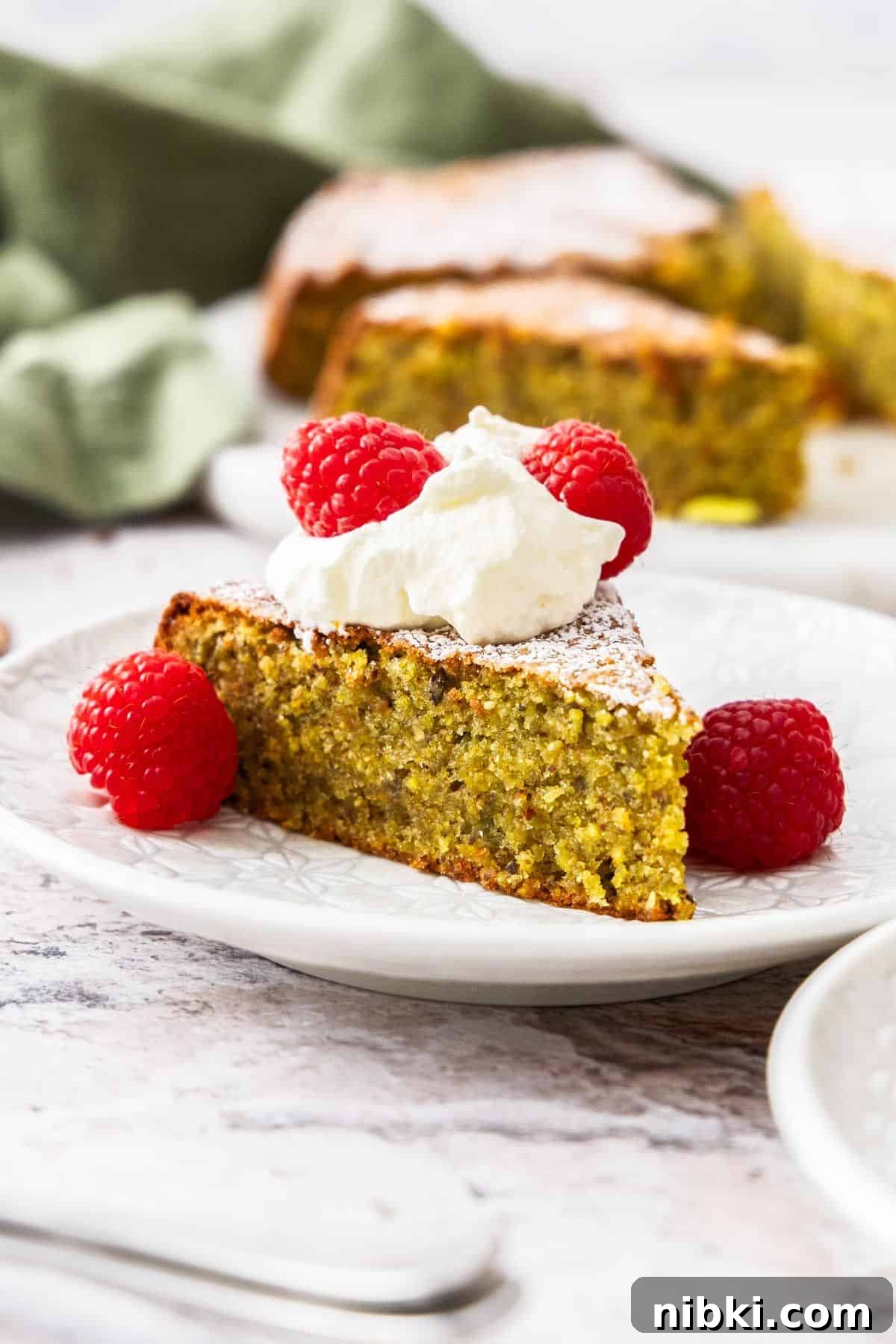 Decadent Flourless Pistachio Cake Recipe: Gluten-Free Dessert 2 Piece of Pistachio Cake on a small white plate, topped with whipped cream and raspberries.