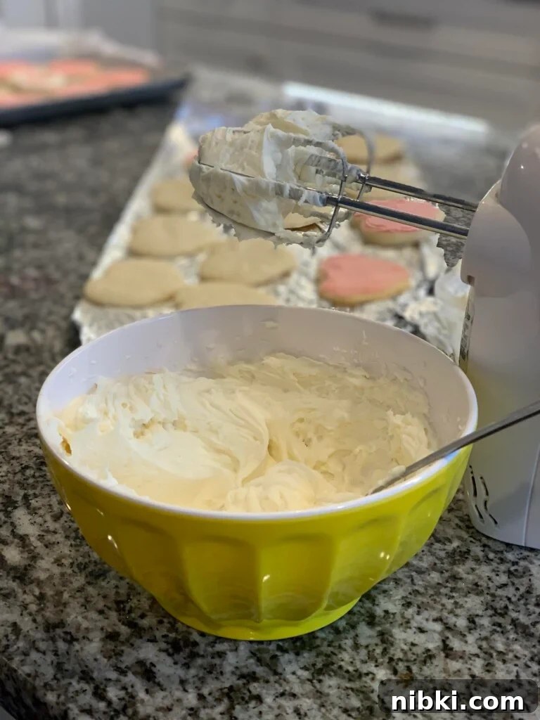 No-Chill Soft Sugar Cookies That Stay Tender and Chewy 12 Frosting in a mixing bowl sits next to a hand mixer and a tray of partially frosted cookies.