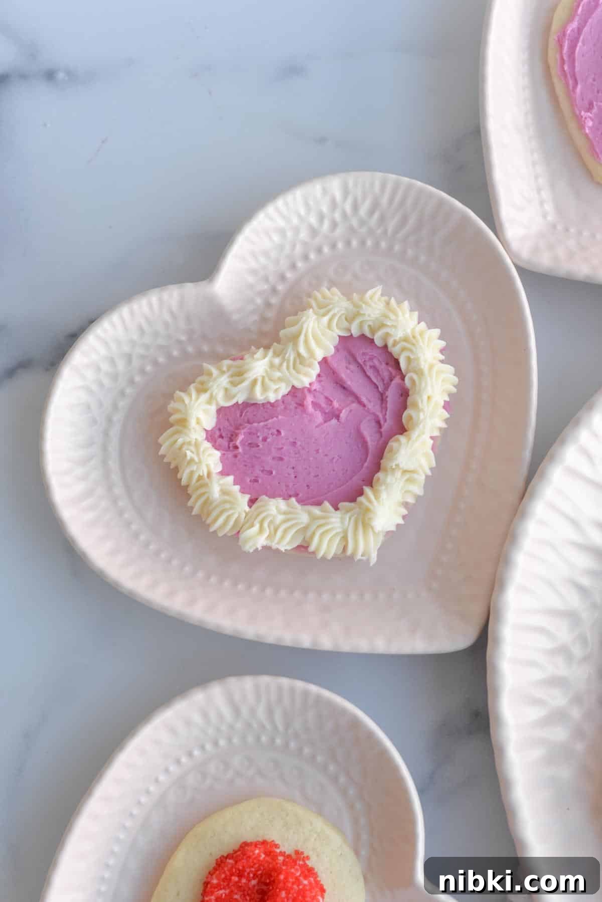No-Chill Soft Sugar Cookies That Stay Tender and Chewy 14 A heart shaped sugar cookie is frosted and sits on a white heart shaped plate.