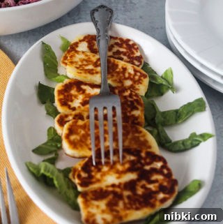 Crispy Grilled or Pan-Fried Halloumi Recipe 6 slices of grilled halloumi cheese on a platter of lettuce with a cabbage coleslaw salad, a stack of plates, and a yellow tablecloth next to it