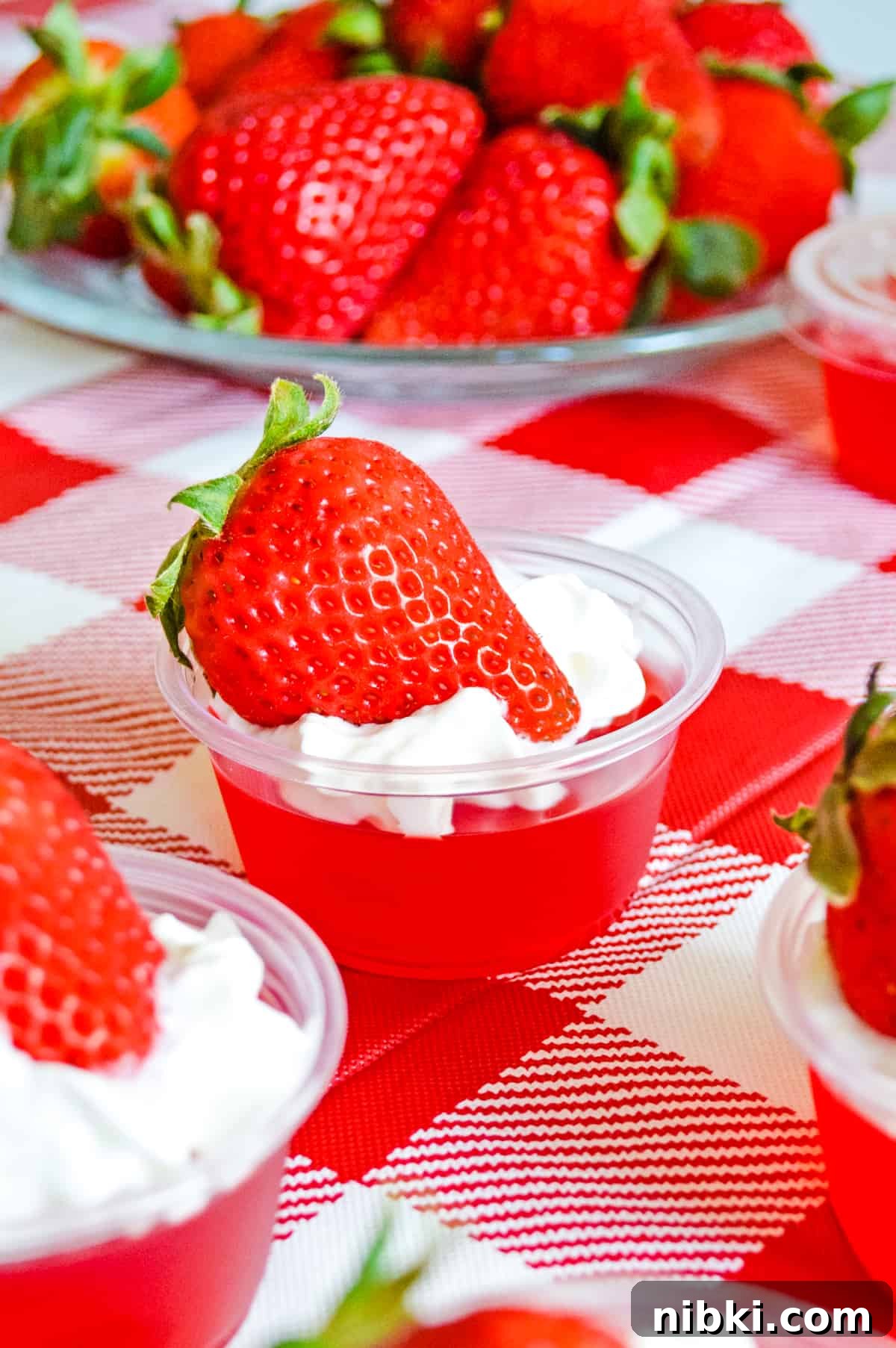 How to Make Strawberry Jello Shots for Parties 2 strawberry jello shots on a red checked tablecloth with a plate of fresh strawberries in the background