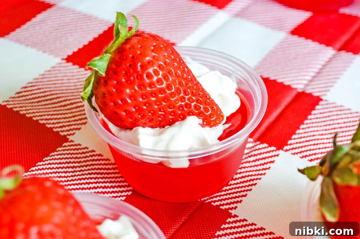 How to Make Strawberry Jello Shots for Parties 3 strawberry jello shot topped with whipped cream and a fresh strawberry, served in a plastic cup and on top of a red and white checkered tablecloth