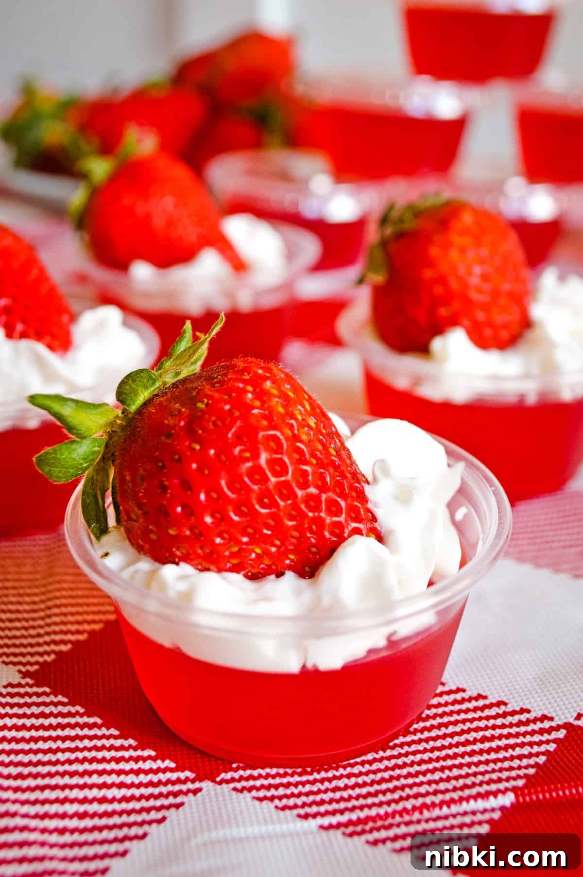 How to Make Strawberry Jello Shots for Parties 5 Strawberry jello shots with whipped cream and strawberry on top, served in plastic shot glasses.