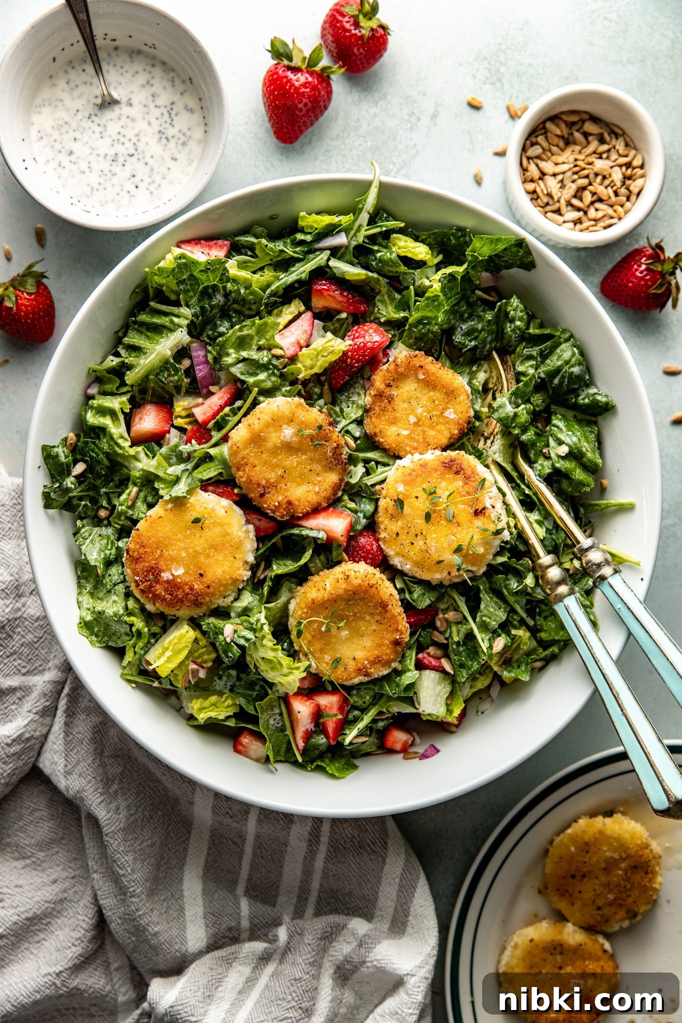 Strawberry Poppyseed Salad with Goat Cheese Croutons 2 The finished strawberry poppyseed salad topped with golden, pan-fried goat cheese medallions, ready to serve in a white bowl with vintage-style utensils.