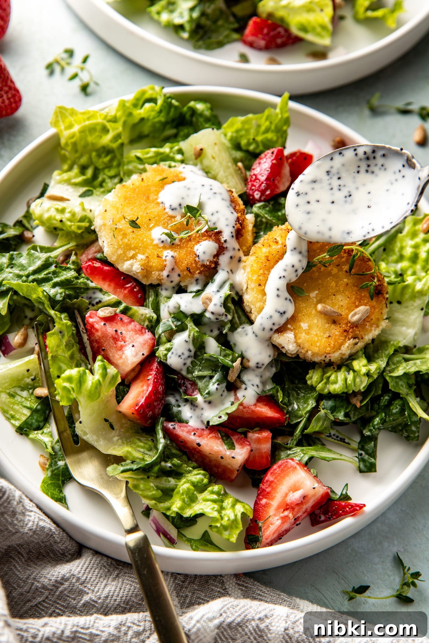 Strawberry Poppyseed Salad with Goat Cheese Croutons 4 Overhead view of a close-up salad plate with two goat cheese medallions, fresh strawberries, and greens, as a spoon pours creamy poppyseed dressing on top.