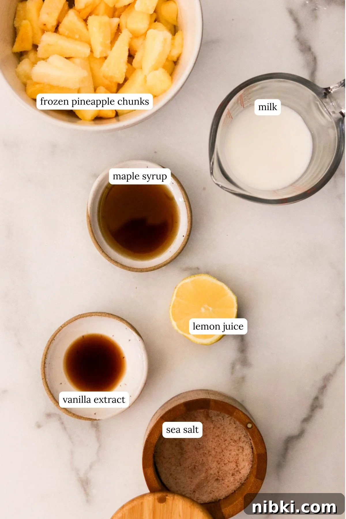 Copycat Disney Pineapple Dole Whip Recipe 2 Ingredients for homemade Dole Whip laid out on a white surface: frozen pineapple, almond milk, maple syrup, vanilla, lemon, sea salt