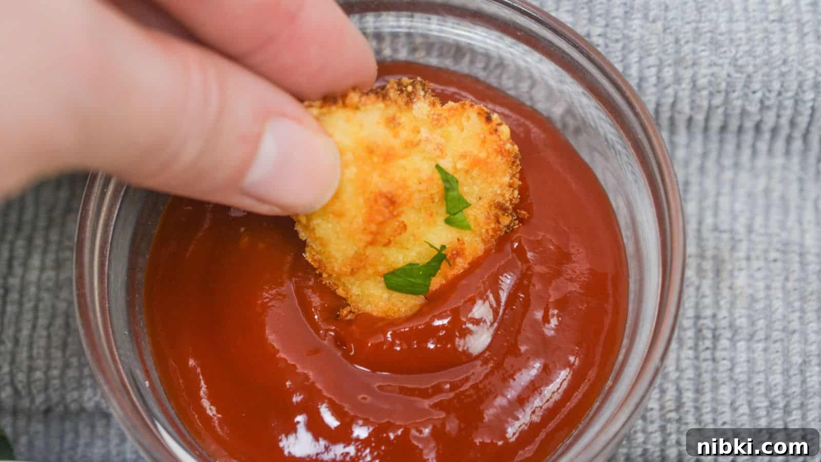 17 Affordable Chicken Recipes That Stretch Your Grocery Budget 14 Hand dipping a chicken nugget into a bowl of ketchup, with a gray cloth background.