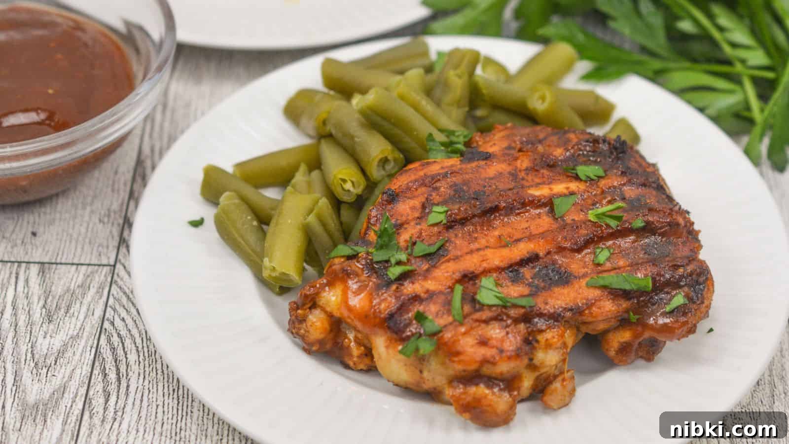 17 Affordable Chicken Recipes That Stretch Your Grocery Budget 4 Grilled chicken thigh on a white plate with green beans and a side of barbecue sauce, garnished with chopped herbs on a wooden table.
