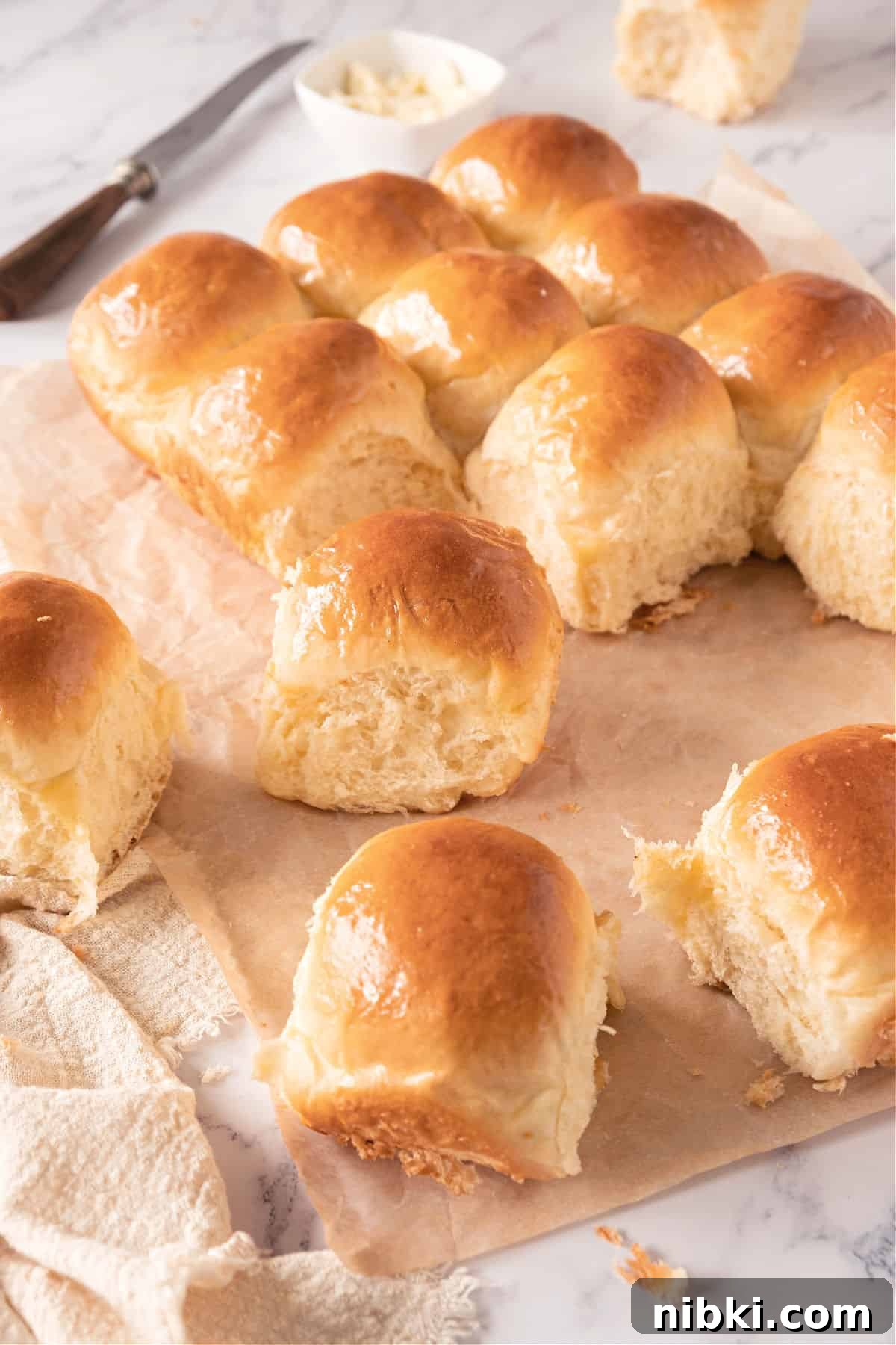 Soft Homemade Yeasted Dinner Rolls 13 Several dinner rolls on beige paper. White marble surface, a knife, kitchen towel.