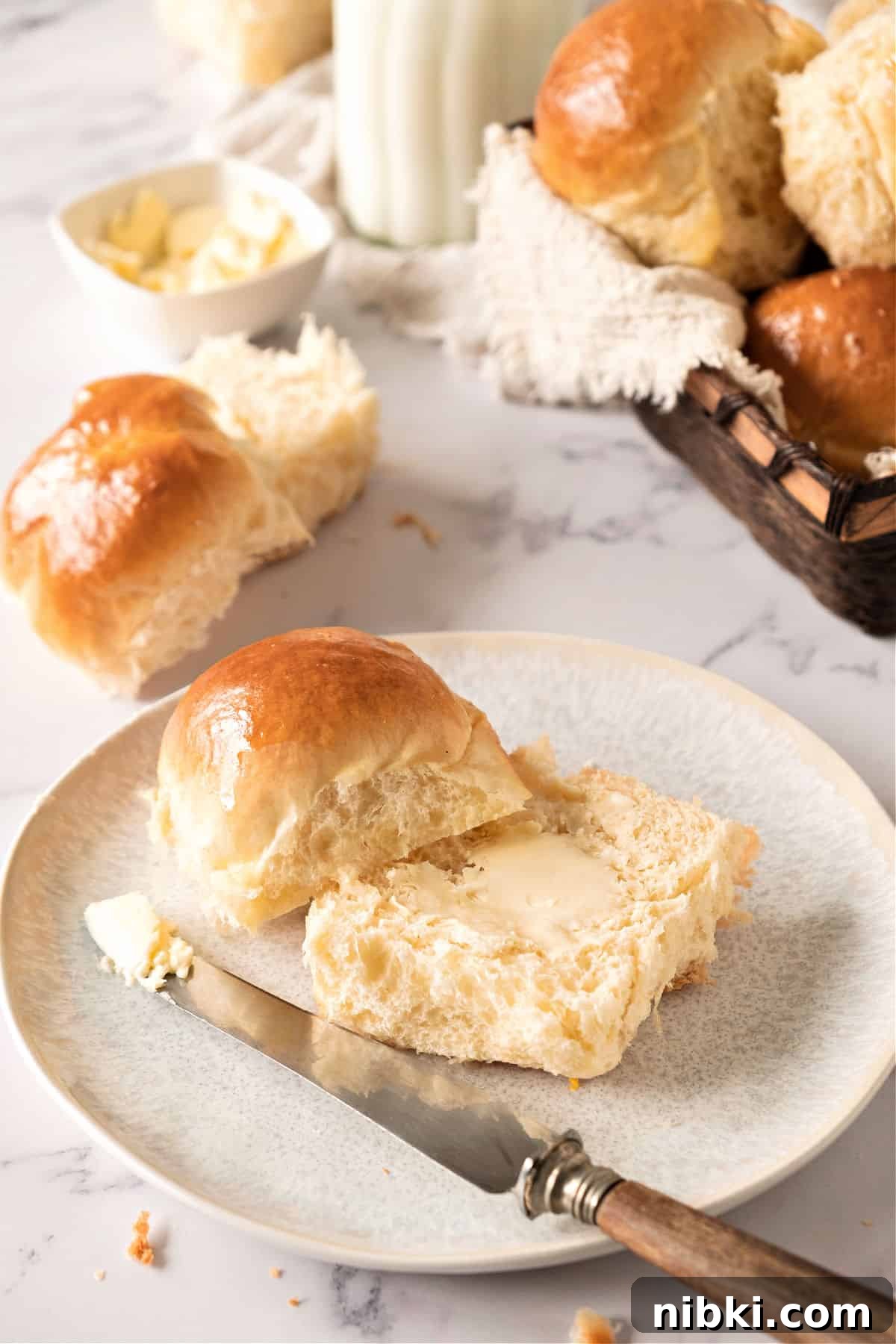Soft Homemade Yeasted Dinner Rolls 3 Halved soft roll with butter. White plate, a knife. White marble surface with more rolls.