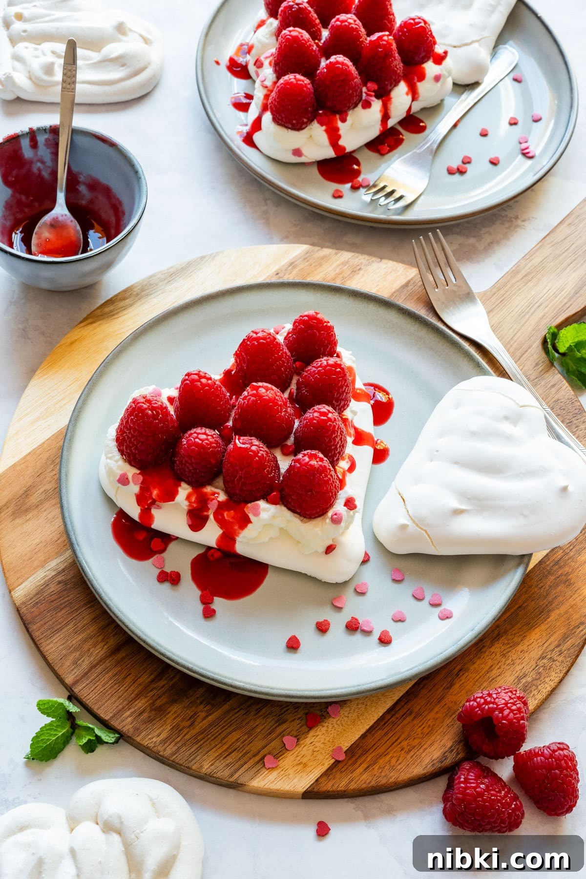Heart-shaped mini pavlovas with whipped cream, fresh raspberries and raspberry coulis on plates.