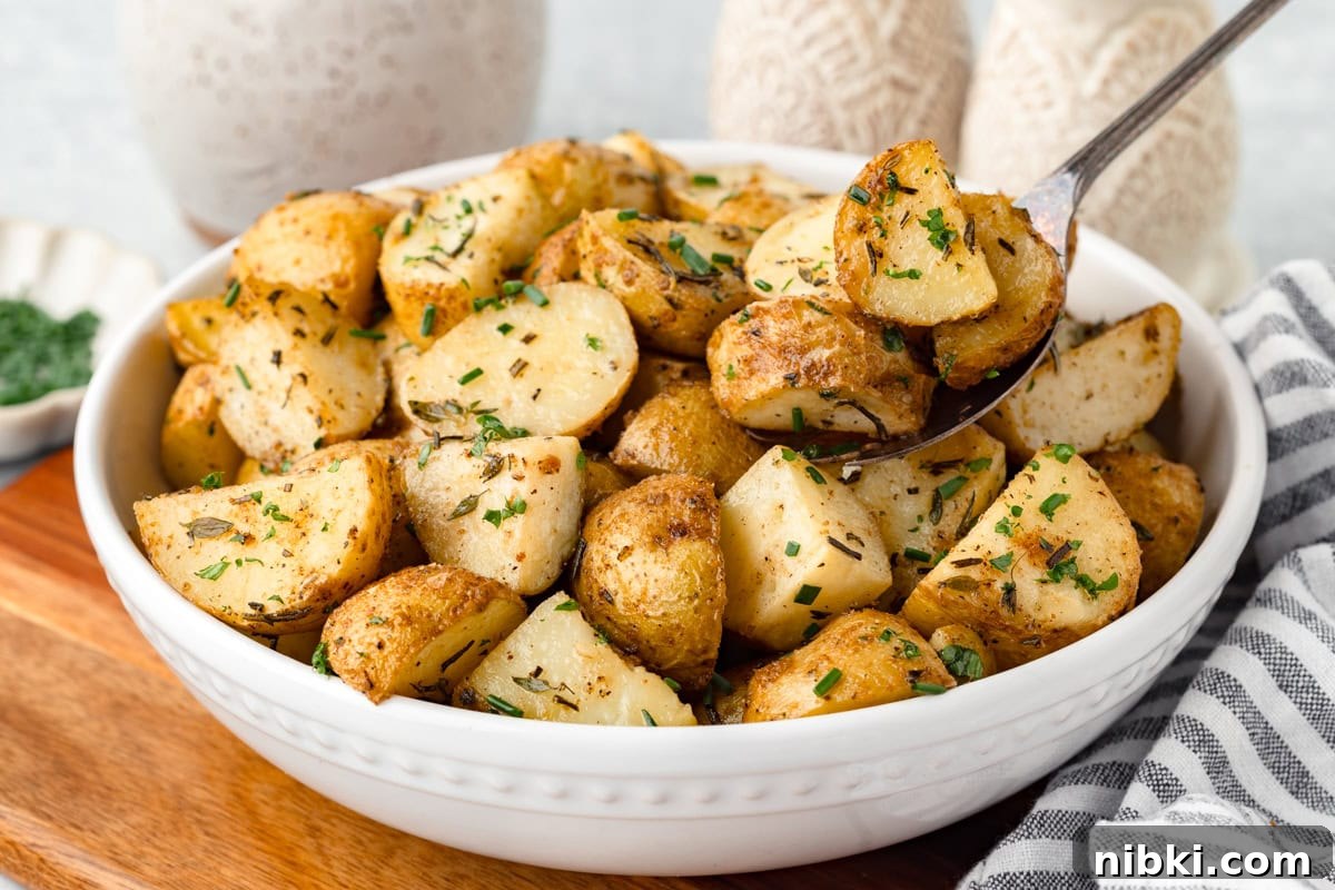A white bowl filled with roasted potato wedges garnished with chopped herbs. A spoon lifts some potatoes from the bowl. The bowl sits on a wooden surface next to a striped cloth napkin.